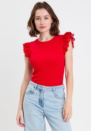 Giovane donna che indossa una canotta rossa senza maniche con spalle in pizzo traforato arricciato e jeans a vita alta di colore azzurro chiaro, in piedi davanti a uno sfondo uniforme.