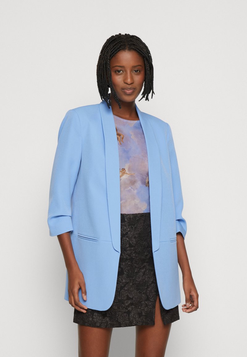 Ljusblå blazer med en skräddarsydd design, med trekvartslånga ärmar och en enknappsstängning. Sammanställd med en blommönstrad topp och svart kjol.