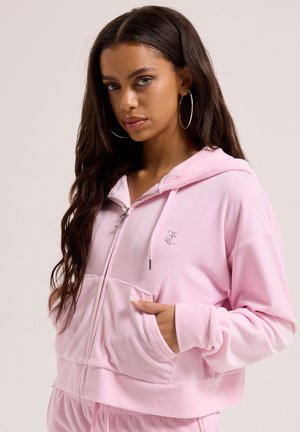 Frau mit langen dunklen Haaren, die Creolen trägt und eine hellrosa Kapuzenjacke mit Reißverschluss trägt, die Hände in den vorderen Taschen.