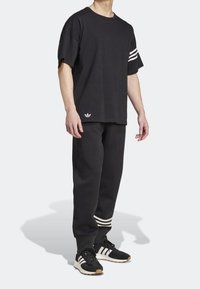 Person klädd i svart Adidas-outfit med vita tre-randsdetaljer på ärmar, byxor och sneakers, stående mot en ljus bakgrund.