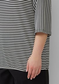Gestreepte T-shirt in zwart en wit, met horizontale strepen, driekwart mouwen en een relaxte pasvorm. Hand rustend op een zwart kledingstuk.