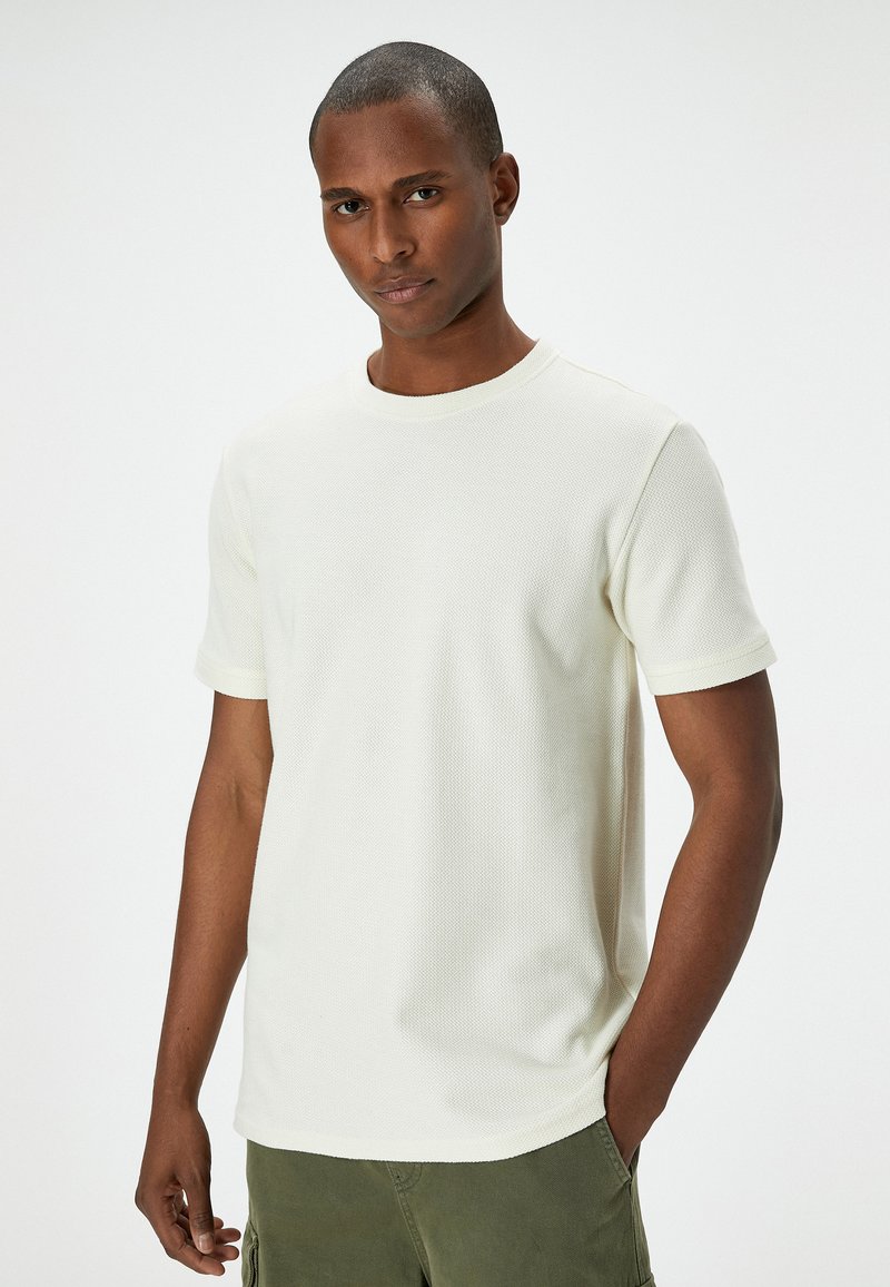 Vit ribbad stickad t-shirt med rund halsringning och korta ärmar, med en slät textur och en avslappnad passform. Matchas med gröna shorts.