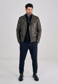 Man staat met handen in de zakken, draagt een donkerbruine leren jas, zwarte trui, zwarte broek en zwarte laarzen, tegen een effen achtergrond.