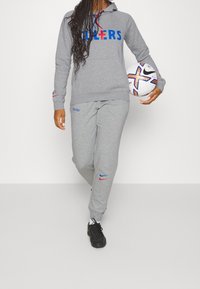 Grauer Hoodie mit blauer und roter Schrift, passende graue Sweatpants und schwarze Sportschuhe. Hält einen weißen Fußball mit bunten Akzenten.