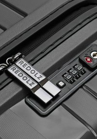 Due zip pull in metallo argentato contrassegnati con "REDOLZ" fissati a una serratura combinata nera su una valigia rigida scura.