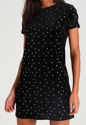 Robe noire à manches courtes avec des petits motifs d'étoiles dorées dispersés, portée par une personne debout les mains relâchées le long du corps.