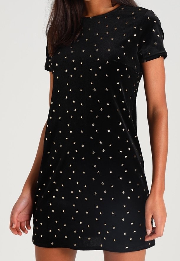 Robe noire à manches courtes avec des petits motifs d'étoiles dorées dispersés, portée par une personne debout les mains relâchées le long du corps.
