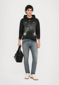 Zwarte hoodie met grafische print, gedragen met lichtblauwe jeans. Accessoires omvatten een zwarte tas en de schoeisel heeft crèmekleurige en bruine accenten.