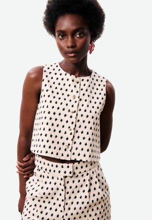 Femme aux cheveux afro portant un haut sans manches blanc et un pantalon avec des taches abstraites noires, ainsi que des boucles d'oreilles en forme de fleur rose.