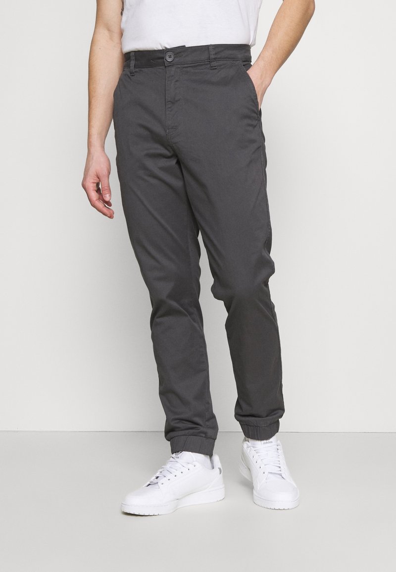 Homme portant un pantalon gris foncé à coupe fuselée avec des bords élastiques et des baskets blanches, debout devant un fond clair uni.