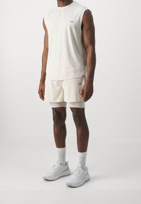 Top senza maniche color beige chiaro e pantaloni corti abbinati, con un logo sottile; abbinati a scarpe da ginnastica bianche e calze. Tessuto dalla trama liscia.