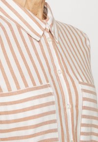Camicia con colletto e bottoni, caratterizzata da righe verticali beige e bianche, con due tasche sul petto e un tessuto morbido e testurizzato.