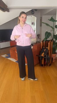 Νέα γυναίκα με ροζ μπουφάν και μαύρο παντελόνι στέκεται στο σαλόνι με ξύλινο πάτωμα και φυτά μέσα στον χώρο.