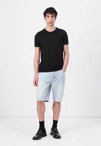 T-shirt en coton noir, shorts en denim bleu clair, chaussettes noires et mocassins noirs. Modèle se tient face à l'avant sur fond blanc.