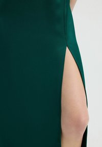 Saténové šaty v smaragdově zelené barvě s bočním rozparkem, který odhaluje pokožku. Hladká textura a elegantní design zvýrazňující siluetu.
