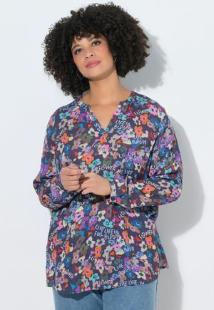 Kobieta o kręconych włosach, ubrana w długą, kwiecistą bluzkę z wielobarwnymi kwiatami oraz napisami "Life in bloom" i "Flower".