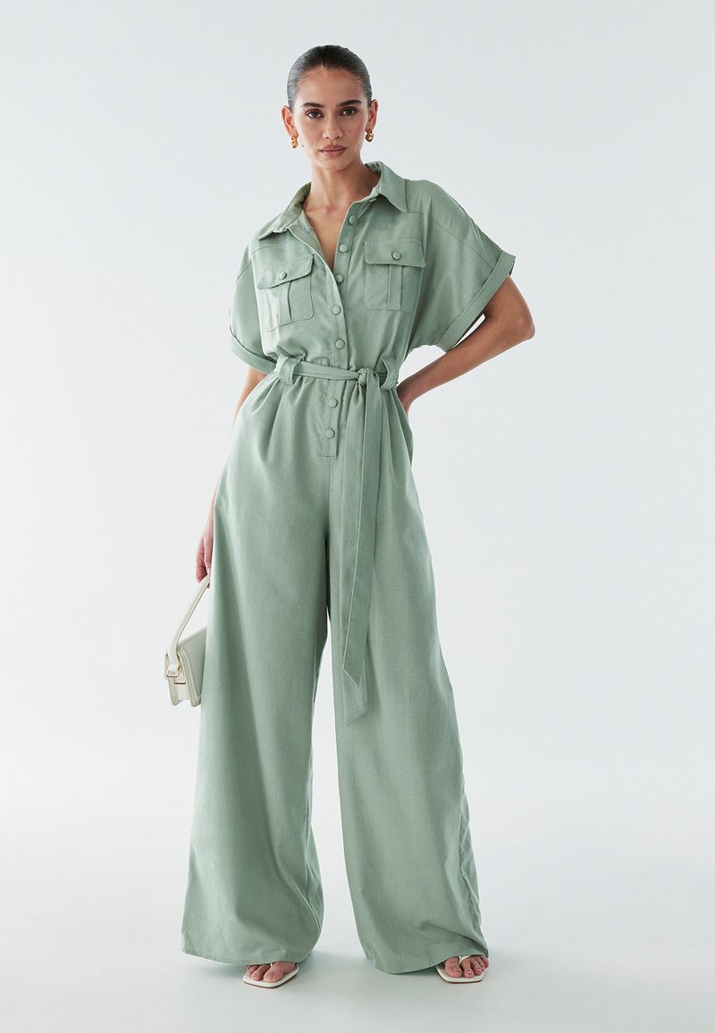 Svetlo zelena kombinezon iz teksturirane tkanine, z kratkimi rokavi, pasom, dvema žepoma na prsih in širokimi hlačnicami.