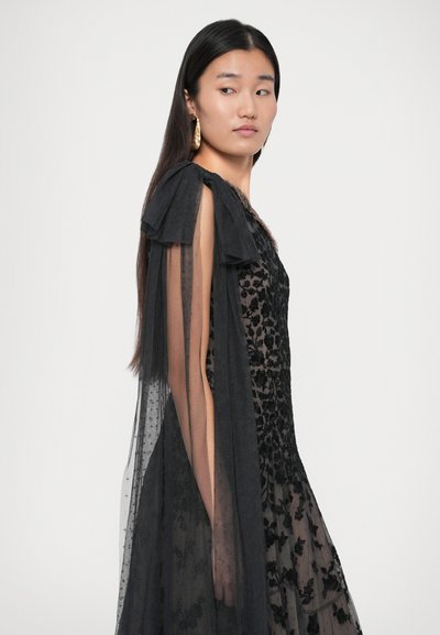 O femeie îmbrăcată într-o rochie transparentă neagră cu modele florale și mâneci lungi din tul, privind peste umăr pe un fundal simplu.