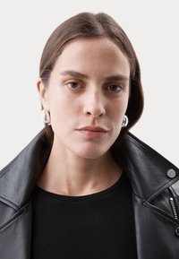 Zwartelerenjack met brede kraag, gecombineerd met een zwart top. Bevat grote zilveren ringoorbellen en een gladde huidtextuur. Neutrale achtergrond.