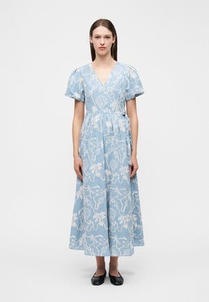 Žena v modrých maxi zavinovacích šatách s bielou výšivkou s oceánovou tematikou a balónovými rukávmi, stojaca vzpriamene v čiernych plochých topánkach na bielom pozadí.