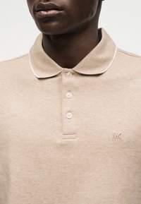 Άνδρας που φοράει μπεζ κοντομάνικη πόλο μπλούζα με τρία κουμπιά και μικρό λογότυπο "MK" στο αριστερό μέρος του στήθους, εμφανίζεται από τον λαιμό μέχρι το μέσο του κορμού.