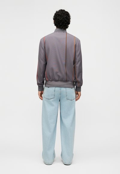 Άτομο με σγουρά μαλλιά στέκεται με την πλάτη, φοράει γκρι σακάκι με κάθετες ρίγες και ανοιχτό μπλε παντελόνι με φαρδιά πόδια.