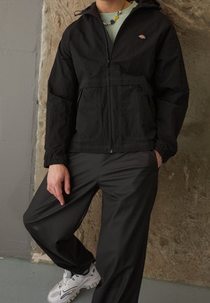 Person trägt eine schwarze Reißverschlussjacke mit Logo, schwarze Hose, weiße Turnschuhe und eine Perlenkette und steht vor einer strukturierten Wand.