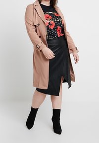 Abrigo trench beige con botones negros, combinado con una camiseta negra con estampado floral y una falda corta de piel sintética negra con una abertura frontal. Botines negros.