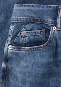 Vaqueros de mezclilla en azul intermedio con costuras visibles. Las características incluyen un pequeño bolsillo frontal y remaches de metal para refuerzo.