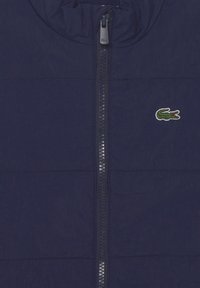 Giacca imbottita blu navy con zip completa, tessuto testurizzato e un piccolo logo di coccodrillo ricamato di colore verde sul petto.