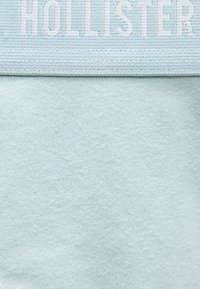 Ανοιχτό μπλε ύφασμα με διπλωμένη άκρη που εμφανίζει μερικώς το όνομα της μάρκας «HOLLISTER» σε μεγάλα, λευκά γράμματα.