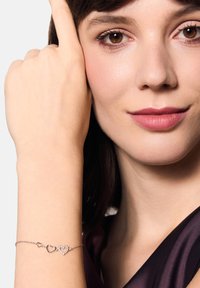 Srebrna bransoletka z sercowo-kształtnymi ogniwami, z mixem gładkich i zdobionych wzorów, noszona na nadgarstku, na neutralnym tle.