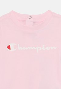 Ružičasta pamučna majica s okruglim izrezom, s bijelim "Champion" logotipom i crvenim akcentom u slovu "C". Stražnje zatvaranje na dugme.
