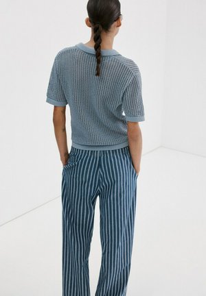 Personne aux cheveux tressés portant un haut à manches courtes en tricot bleu clair et un pantalon rayé bleu marine, debout les mains dans les poches.