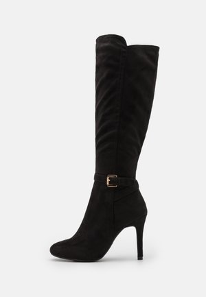 Botte noire montante jusqu’au genou avec un talon aiguille fin et une petite bride à boucle dorée autour de la cheville.