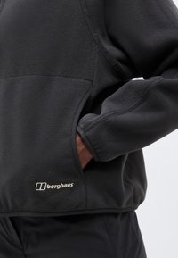 Μαύρο φλις μπουφάν Berghaus με το χέρι στην πλαϊνή τσέπη, ορατές ραφές και κεντημένο λογότυπο της μάρκας κοντά στο τελείωμα.