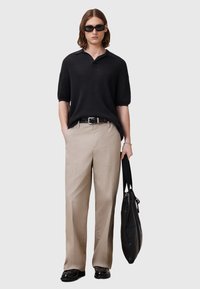 Svart stickad polo skjorta, beige vida byxor, svart bälte och stor svart väska. Man med mörka solglasögon och rakt hår.