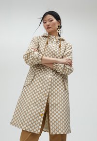 Jasnobeżowy płaszcz w wzory o geometrycznym nadruku, z kapturem ze sznurkiem i guzikami. Posiada elastyczne mankietami i boczne kieszenie.