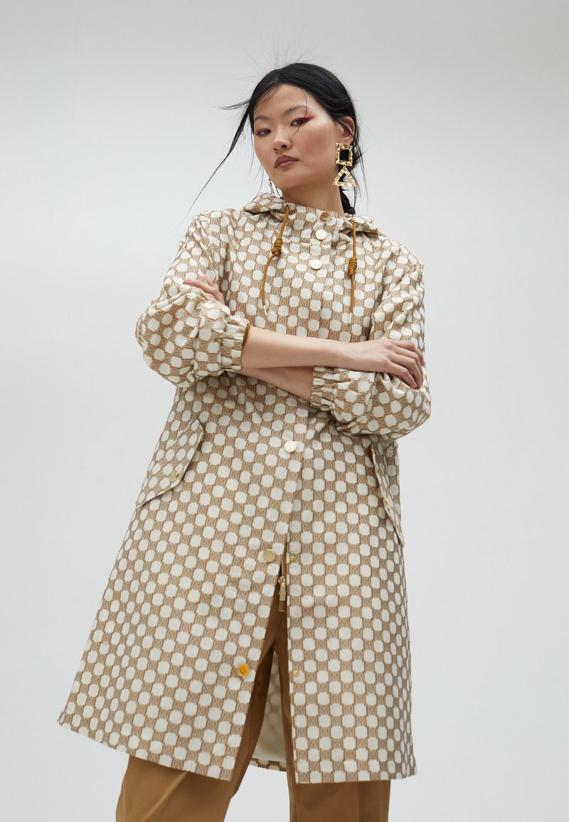 Jasnobeżowy płaszcz w wzory o geometrycznym nadruku, z kapturem ze sznurkiem i guzikami. Posiada elastyczne mankietami i boczne kieszenie.