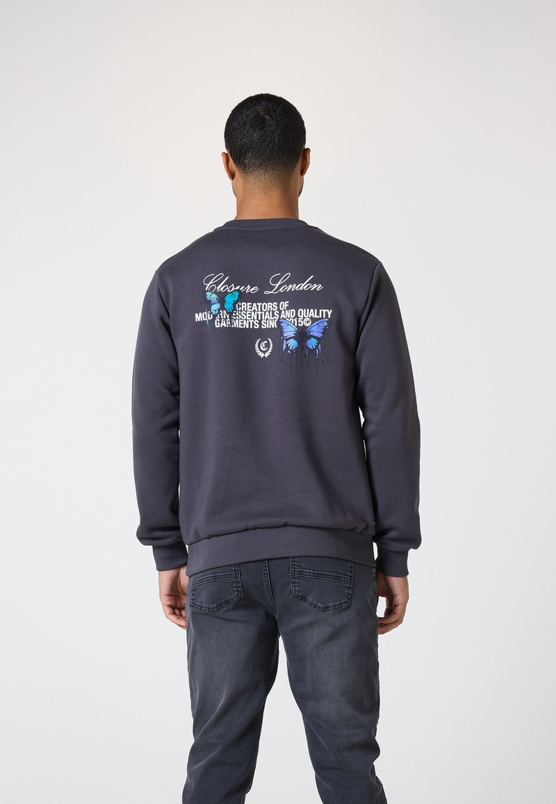Sudadera gris oscuro con puños y dobladillo acanalados. La parte trasera presenta un gráfico de mariposa azul y texto blanco. Combinada con jeans oscuros.