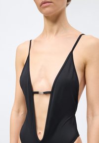 Γυναίκα με μαύρο ολόσωμο μαγιό με λεπτές τιράντες και βαθύ V ντεκολτέ που συγκρατείται από ένα μικρό ασημένιο κούμπωμα στο στήθος.