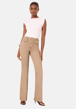 Vrouw staand, dragen beige hooggetailleerde rechte broek en een mouwloos wit topje met gouden knoopdetails en bruine hakken.