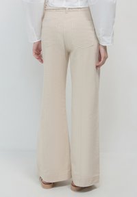 Persona che indossa pantaloni beigi a vita alta e a gamba larga, con una camicia bianca a maniche lunghe, in piedi con le mani rilassate ai lati.