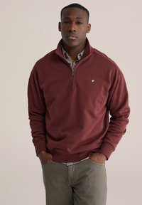 Hnedá mikina na zips z mäkkého materiálu, s rebrovanými manžetami a lemom. Malé logo je umiestnené na ľavej strane hrude. Nosená cez košeľu s golierom.