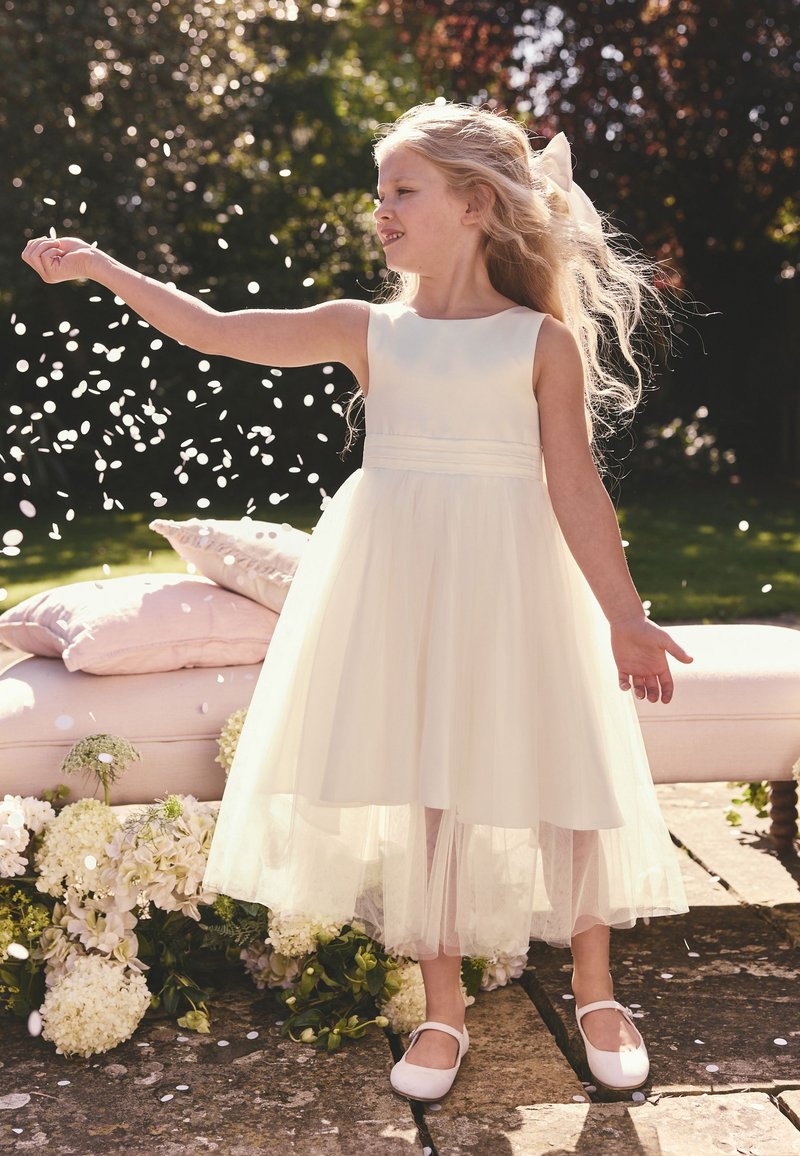 Giovane ragazza in abito bianco che lancia petali di fiori all'aperto vicino ad allestimenti floreali e cuscini morbidi in una giornata di sole.