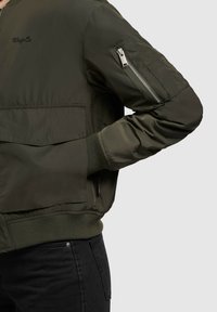 Olijfgroene bomberjack met een geribbelde onderrand, een ritszak op de mouw en een geborduurd logo op de borst. Gemaakt van glad, lichtgewicht materiaal.