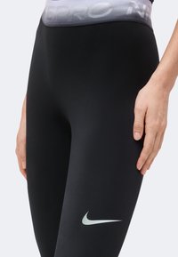 Gros plan sur un legging noir Nike avec une ceinture grise et le logo blanc Nike sur la cuisse, porté par une personne avec la main posée sur la hanche.