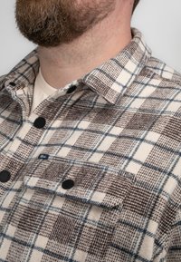 Beżowo-brązowa flanelowa koszula w kratę z czarnymi guzikami, kołnierzykiem oraz kieszenią na piersi z metką marki. Miękka tekstura i zorganizowany design.