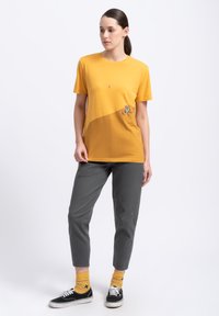 Geel T-shirt met korte mouwen en een gebogen ontwerp, voorzien van een kleine grafische accent aan de voorkant, gecombineerd met grijze tapered broek en zwarte sneakers.