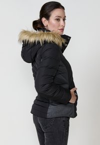 Crna i siva puffer jakna s umetkom od lažnog krzna na kapuljači, kviltana tekstura, džepovi na patentni zatvarač i prilagođeni dizajn.